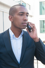 Serious stressed businessman talking on mobile phone outdoors. Pensive African American man in suit calling on cellphone and listening. Phone talk concept