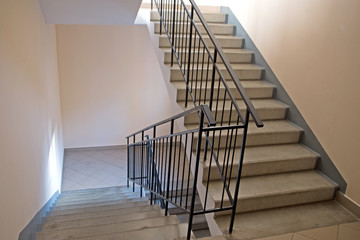 Stairs in the entrance of an apartment building. Two flights of stairs with concrete steps and black metal railings along beige walls, the landings at the bottom is lined with large gray tiles - Powered by Adobe