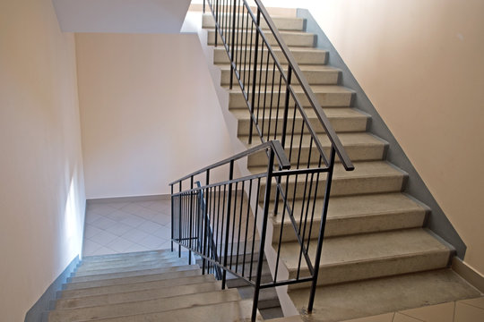 Stairs in the entrance of an apartment building. Two flights of stairs with concrete steps and black metal railings along beige walls, the landings at the bottom is lined with large gray tiles