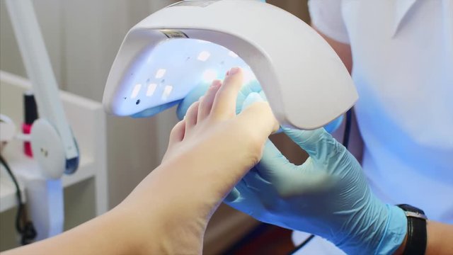 Professional pedicure is beauty salon. Pedicurist woman in gloves is holding UV lamp above the foot. She is drying shellac on client toes, foot closeup.