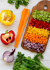 Chopped fresh vegetables (carrot, celery, red onion, peppers) arranged on a cutting board on a white wooden background, top view. From above, overhead, flat lay. Close-up.