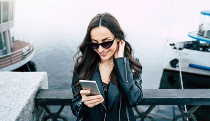 Stylish and trendy young beautiful woman in sunglasses and in hipster style wear while she is using a phone and have a fun outdoors in city. 