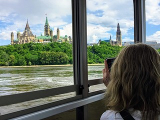Tourists taking in the views of Parliament Hill and the cityscape while taking a tour boat across the Ottawa River from Gatineau to Ottawa on a beautiful day
