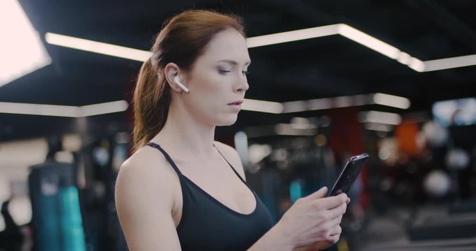 Gym, woman athlete in wireless headphones walks on a treadmill and uses smartphone, tracking progress in sports, aerobic exercise and endurance training.