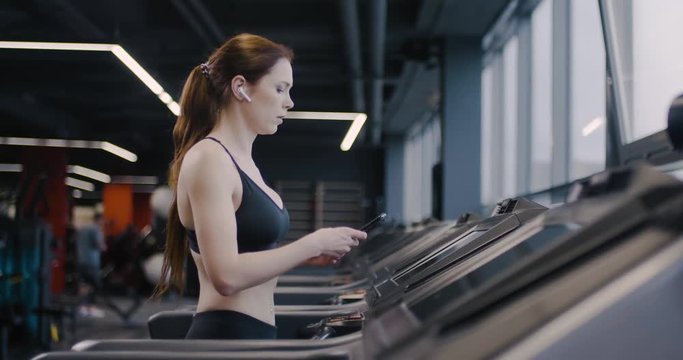 Gym, woman athlete in wireless headphones walks on a treadmill and uses smartphone, tracking progress in sports, aerobic exercise and endurance training.
