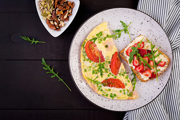 Omelette with tomatoes, ham, green onion and sandwich with strawberry on dark table.  Frittata - italian omelet. Top view