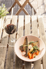 Fried mackerel fish with vegetable garnish: spinach in coconut milk, radish, onion and baked tomatoes, served on a sunny terrace with a glass of red wine