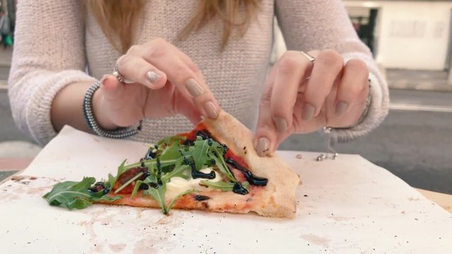 Slow motion shot of a woman with jewelry and painted fingernails takes one slice of arugula pizza with balsamic sauce in outdoor restaurant near street's traffic