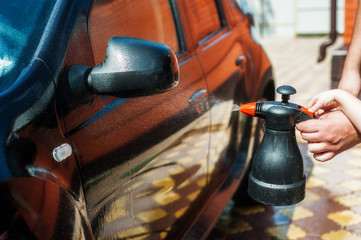 A man sprays car shampoo on a black car from a plastic bottle.