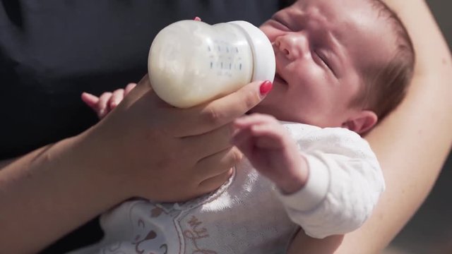 Young beautiful mother is feeding her little baby with a bottle of a milk, but baby is crying.