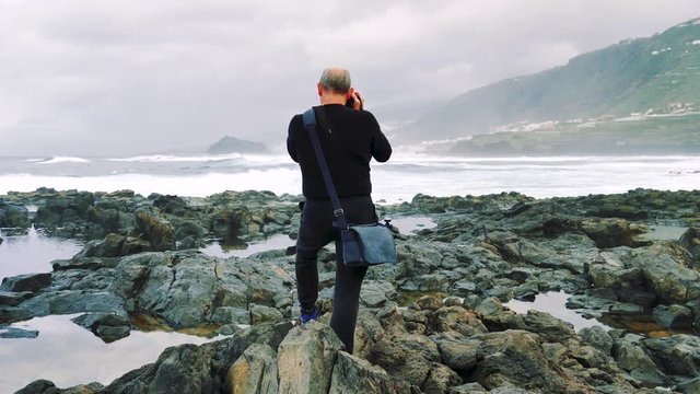 Photographer stands on the rocky coast of Tenerife taking pictures of the surf and landscape on a gloomy overcast day. Beautiful winter coastal scenery of the Canary Islands. Slow motion.