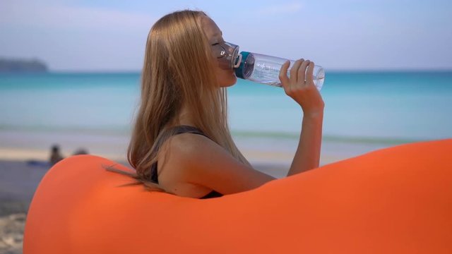 Superslowmotion shot of a young woman on a tropical beach sits on an inflatable sofa and drinks water from a multi useable plastic bottle. Drink more water. Reusable plastic and Reduce plastic waste