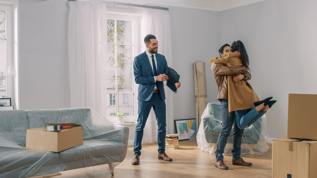 Real Estate Agent Shows Bright New Apartment to a Young Couple. Successful Young Couple Becoming Homeowners. Girl Jumps Into His Boyfriend's Arms Hug. Spacious Bright Home with Big Windows.