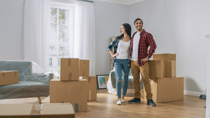 Happy Young Couple Moving Into New Apartment, Carrying Cardboard Boxes with Stuff and Poses on Camera. Young Boyfriend and Girlfriend Start Living Together, Unpacking Stuff.