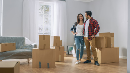 Happy Young Couple Moving Into New Apartment, Carrying Cardboard Boxes with Stuff and Poses on Camera. Young Boyfriend and Girlfriend Start Living Together, Unpacking Stuff.