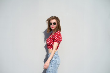 Redhead caucasian girl in red polka-dot pattern blouse and jeans staying near grey background