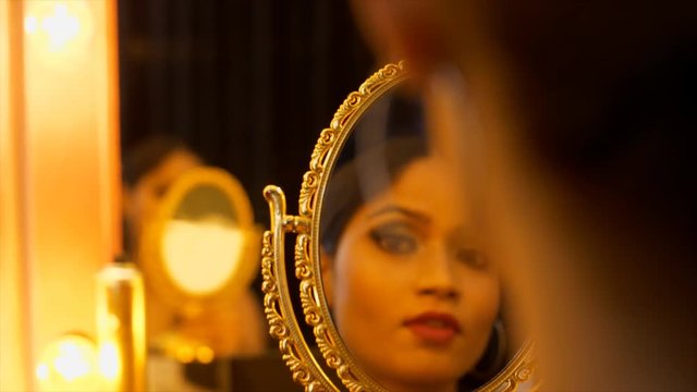 The young lady is done with her party makeup and checking her styling in a golden framed mirror . The young beautiful girl looking at the mirror to check her makeup and styling - Makeup studio 