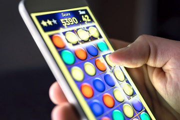 Mobile game in smartphone screen. Young man playing puzzle game in phone app. Gamer having fun. Digital gaming, entertainment and videogame concept. Lazy or addicted man late at night.