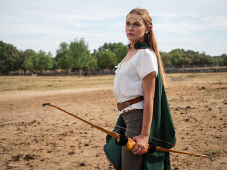 Blonde girl with elf ears poses in the field with a bow and a green cape.