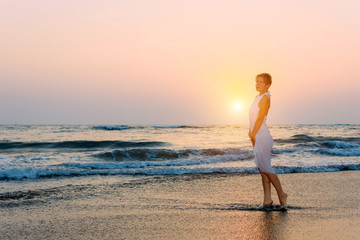 Fototapeta na wymiar Attractive slender girl in white dress stands in waves on the sea in the light of setting sun. Young woman walks barefoot along the surf and looks into the distance. Rough sea and ripples on water.