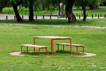 Public park wooden table with benches supported with metal frame surrounded with stone tiles uncut grass and trees in background on warm sunny spring day