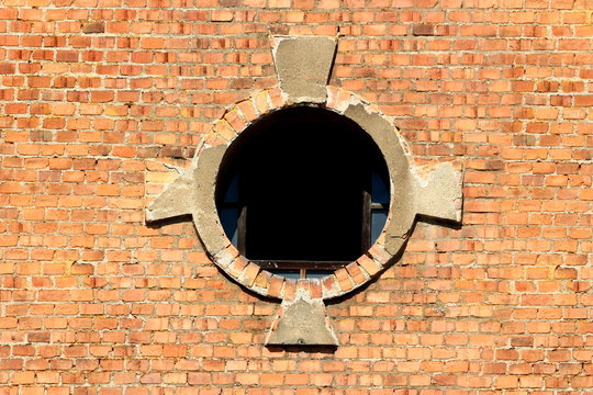Round window opening with partially broken glass and decorative red bricks tiles frame on old abandoned industrial complex building on warm sunny spring day © hecos