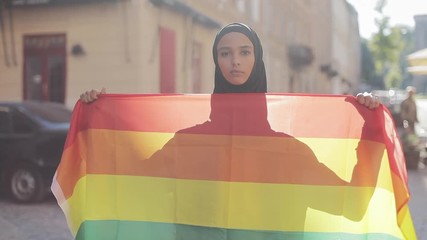 Portrait of beautiful young Muslim woman wearing hijab headscarf with LGBT flag standing on the old street background. Lesbianism and Islam concept.