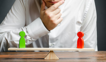 Man holds hand on chin looks at the rival red and green figures on a scales. Weighted decision. The...