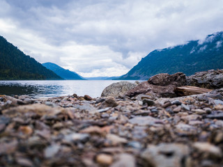 Close-up on the round stones of gray-brown color on the shore of the island against the backdrop of Teletskoye lake or a large river that goes into the distance, meandering between mountains of Altai