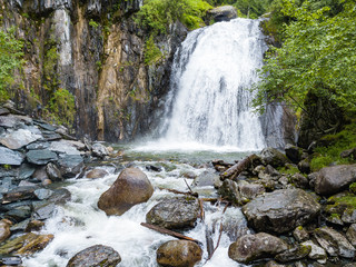 A large waterfall in the back of the Altai Mountains with gray-brown stones near a steep cliff with green trees. Rest and loneliness while traveling to deserted places.