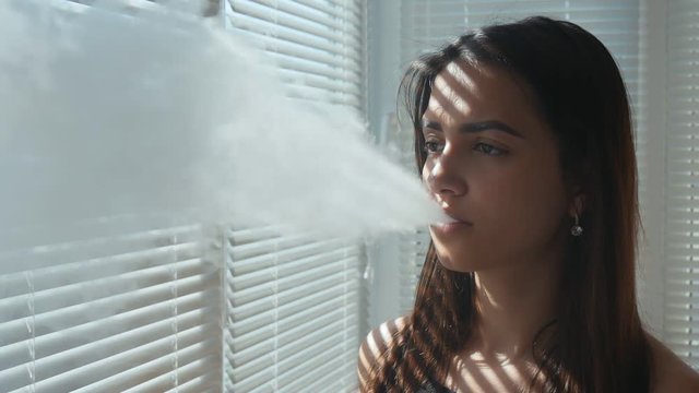 Smiling young brunette woman vaping on the balcony in the office with jalousie on the windows with sun rays on the face, Slow motion