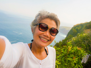 middle age 50s happy and cheerful Asian woman with grey hair taking selfie with mobile phone at beautiful tropical beach island smiling at cliff viewpoint enjoying Summer