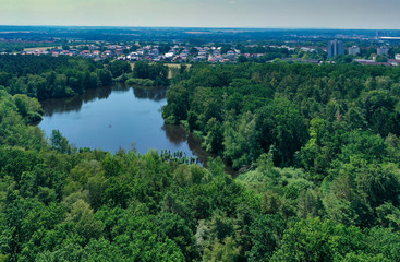 Fototapeta na wymiar Aerial view of a densely overgrown green forest area with a large old pond and the houses of the city in the background.