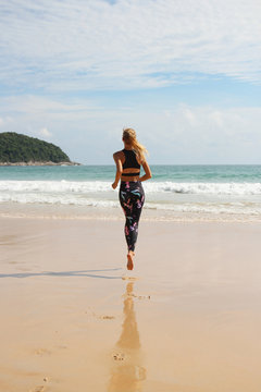 Back view: slim blonde girl wearing black tracksuit with the image of sea horses runs to the sea. Concept of sports life