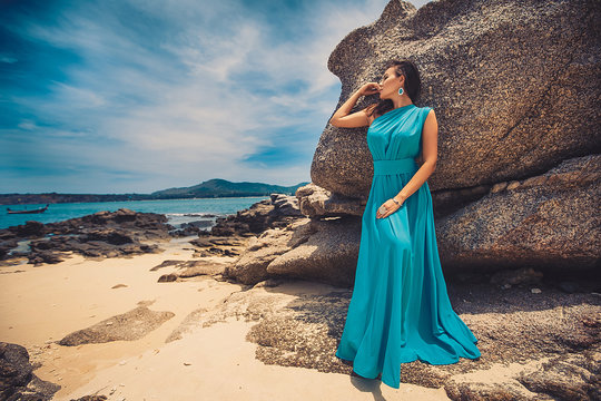 Sexy girl in a blue long dress posing near rock on beach with white sand. Woman resting and sunbathing on paradise island.