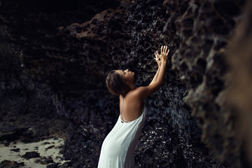 Beautiful sensual woman wearing long white dress and posing beside the black cliffs and touching their. Mysterious black caves.