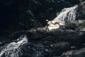 Young beautiful woman in white dress is posing at waterfall