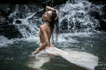 Fototapeta premium Close up photo of Young beautiful woman with nude back in white dress is standing in the water on a big waterfall.