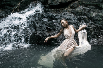 Fashion model girl in a white long dress against a waterfall background. Dark background.