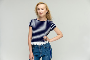 Portrait of a pretty interesting blonde girl on a white background in jeans and a striped blue t-shirt. Standing in front of the camera, smiling, showing hands. Shows a lot of emotions.
