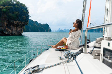 A beautiful asian lady in a white shirt on a yacht drinks champagne and eats fruit, on the background of sea. Back  view - obrazy, fototapety, plakaty