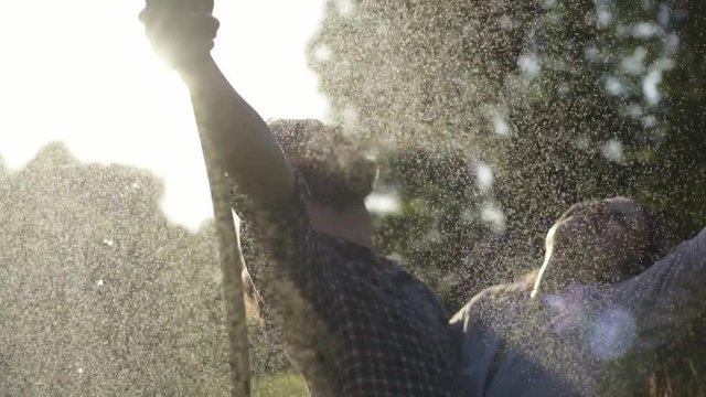 Young cheerful beautiful couple hosing the garden and playing under sprays of water at the sunset time. Summer happiness and love concept