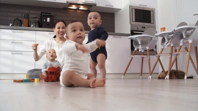 Full shot of happy middle-aged Asian woman sitting on floor in kitchen, singing to music and snapping fingers, and adorable 1-year-old toddler triplets, one standing and others sitting, waving arms