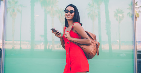 Half length portrait of cheerful woman in fashionable sunglasses holding mobile phone for chatting in hands and laughing at camera during day for walking, gladden jubilate hipster girl phoning