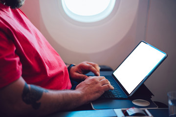 Young man flight passenger connecting to wireless internet on board with touch pad while sitting next to aircraft cabin window. Mock up blank screen for web template, digital nomad work on freelance