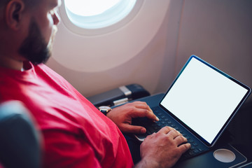 Young man flight passenger connecting to wireless internet on board with touch pad while sitting next to aircraft cabin window. Mock up blank screen for web template, digital nomad work on freelance