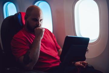 Young man flight passenger watching movie on board via entertainment Tv while sitting in comfortable airplane seat next to aircraft cabin window. Caucasian man enjoying travel via business class