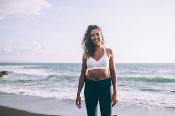 Portrait of cheerful young woman dressed in active wear happy from morning training on coastline while smiling at camera, positive dark skinned female in sport clothes standing on sea shore