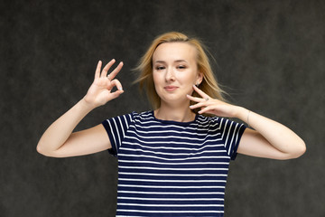 Portrait to the waist of a pretty interesting fashionable blonde girl on a gray background in a striped blue t-shirt. Standing in front of the camera, smiling, showing hands. Shows a lot of emotions.