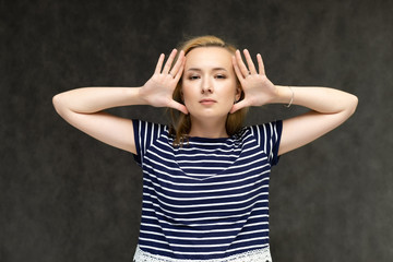 Portrait to the waist of a pretty interesting fashionable blonde girl on a gray background in a striped blue t-shirt. Standing in front of the camera, smiling, showing hands. Shows a lot of emotions.